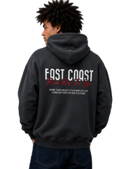 A man wearing a dark charcoal gray hoodie from the back, featuring 'FAST COAST' and 'Kean Keen Tee Co.' printed in white and red on the back, paired with dark blue jeans against a solid white background.