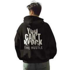 A person viewed from behind wearing a black hoodie with large white text reading 'YOU CAN'T KNOCK THE HUSTLE' on the back, standing on a city street.