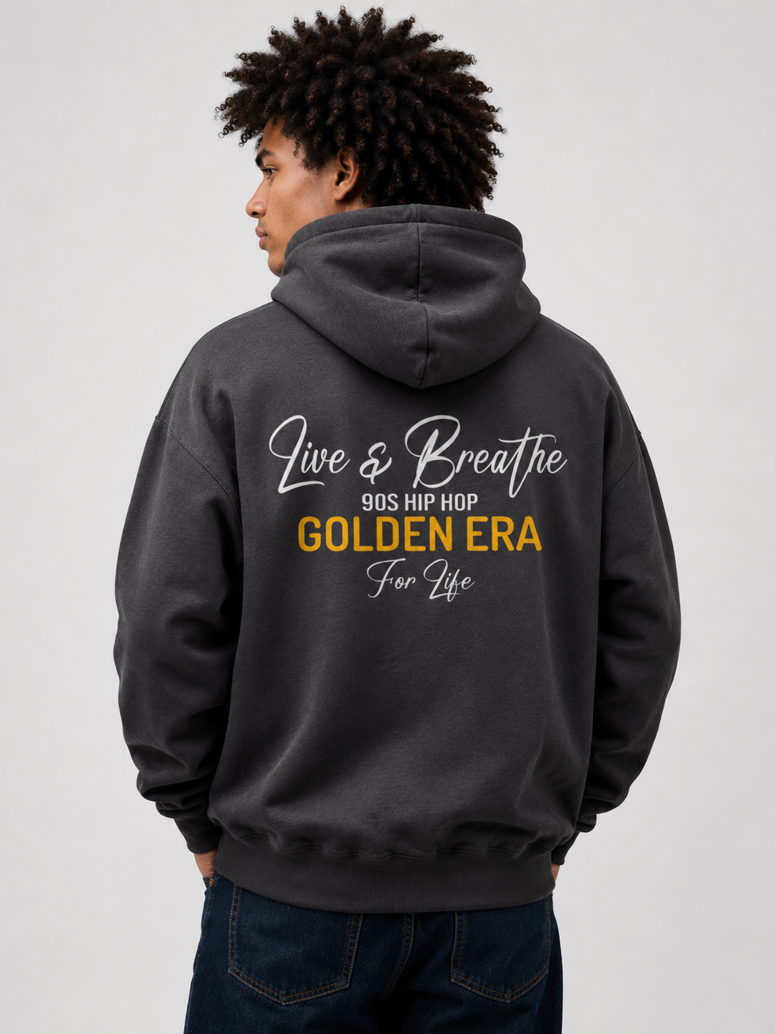 A man wearing a dark gray hoodie from the back, featuring a graphic print that reads 'Live & Breathe 90s Hip Hop Golden Era For Life' in white and yellow text. He is also wearing dark blue jeans against a plain light gray background.