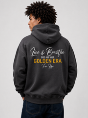 A man wearing a dark gray hoodie from the back, featuring a graphic print that reads 'Live & Breathe 90s Hip Hop Golden Era For Life' in white and yellow text. He is also wearing dark blue jeans against a plain light gray background.