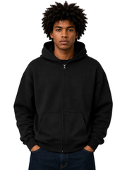 A man with an afro hairstyle wearing a black full-zip hoodie and dark blue jeans, standing against a neutral gray studio background.