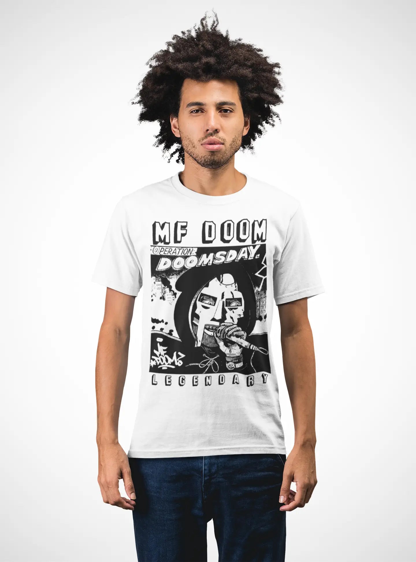 A man with an afro hairstyle wearing a white graphic t-shirt featuring a black and white print of MF Doom and the text 'MF DOOM REIGNITE DOOMSDAY LEGENDARY', paired with dark blue jeans, against a plain white background.