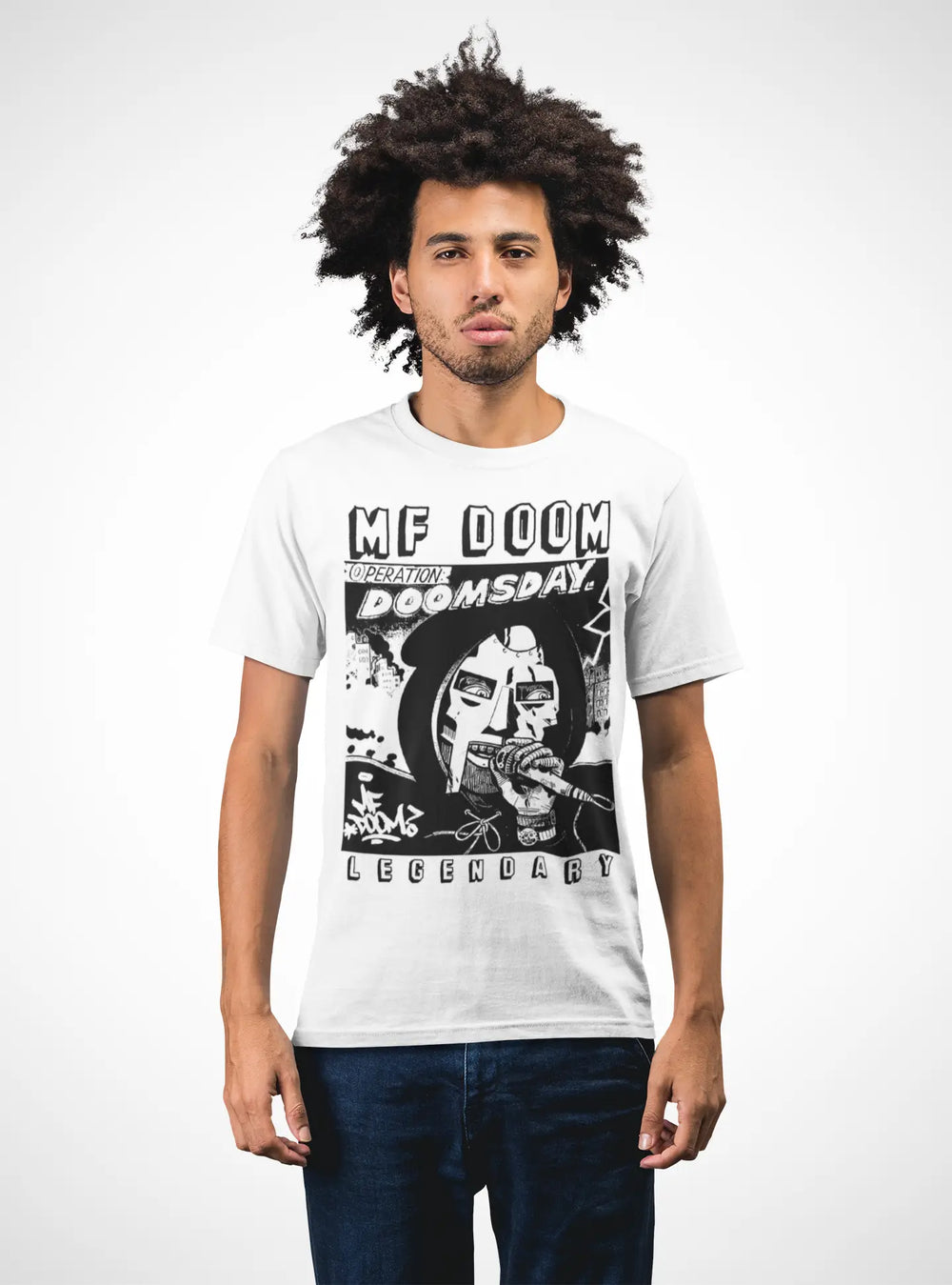 A man with an afro hairstyle wearing a white graphic t-shirt featuring a black and white print of MF Doom and the text 'MF DOOM REIGNITE DOOMSDAY LEGENDARY', paired with dark blue jeans, against a plain white background.